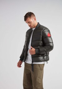 Giacca a vento nera con design bomber, colletto a coste, dettaglio patch rosso e chiusura con zip. Indossata sopra una camicia bianca e pantaloni oliva.