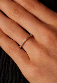 Gold band ring with small, clear gemstones set in a row, worn on a hand against a dark textured background. Smooth and polished finish.