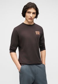 Νεαρός άνδρας με μεσαίου μήκους σκούρα μαλλιά, φορώντας μαύρο μακρυμάνικο πουκάμισο με πορτοκαλί κείμενο, με τα χέρια στις τσέπες, μπροστά σε απλό ανοιχτό φόντο.