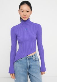 Maglione a collo alto viola con coste, caratterizzato da maniche lunghe con estremità svasate, orlo asimmetrico e una toppa blu sul petto. Abbinato a jeans azzurri chiari.