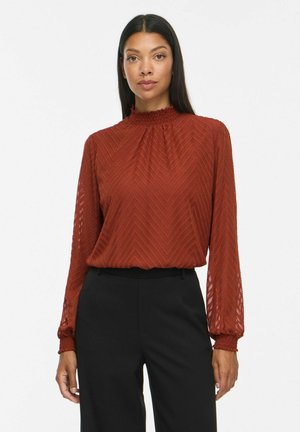 Femme portant un chemisier texturé manches longues de couleur rouille avec un col montant et un pantalon noir sur un fond uni.