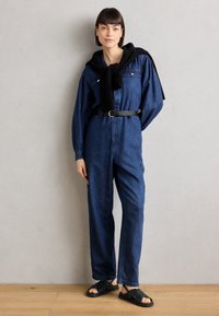 Blå denim jumpsuit med lange ermer, brystlommer og innsnørt liv. En svart genser er drapert over skuldrene. Sorte sandaler fullfører looken.