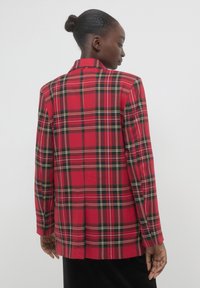 Blazer a scacchi rossi con accenti neri e gialli. Vestibilità sartoriale, maniche lunghe e colletto a scollo a V. La parte posteriore presenta uno spacco centrale per agevolare i movimenti.