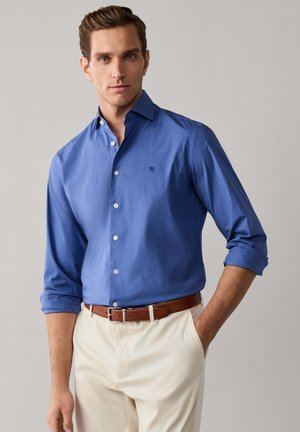 Hombre con camisa azul de botones con mangas remangadas, pantalones beige y cinturón marrón, de pie con una mano en el bolsillo frente a un fondo gris.