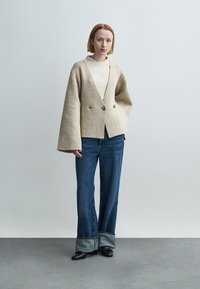 Γυναίκα με κοντά ξανθά μαλλιά που φοράει μπεζ φαρδιά ζακέτα, λευκή μπλούζα, τζιν με ρεβέρ και μαύρα παπούτσια, στέκεται σε γκρι δάπεδο.