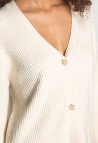 Rippstrick-Cremekardigan mit V-Ausschnitt und zwei Knöpfen. Weiche Textur mit lockerer Passform, die den Stretch des Stoffs hervorhebt.