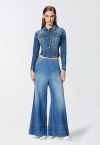 Izrezana traper jakna s kopčanjem na dugmad, uparena sa širokim plavim traper hlačama sa ukrasnim rubom. Oboje imaju izblijeđenu teksturu.