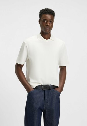 Weißes Poloshirt mit kurzen Ärmeln und V-Kragen, kombiniert mit dunklen Jeans. Der Stoff wirkt glatt und leicht. Schwarzer Gürtel mit Logo.