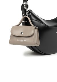 Pochette mini beige in pelle Lancaster Paris attaccata alla tracolla di una borsa a mano nera in pelle curvata su sfondo bianco.