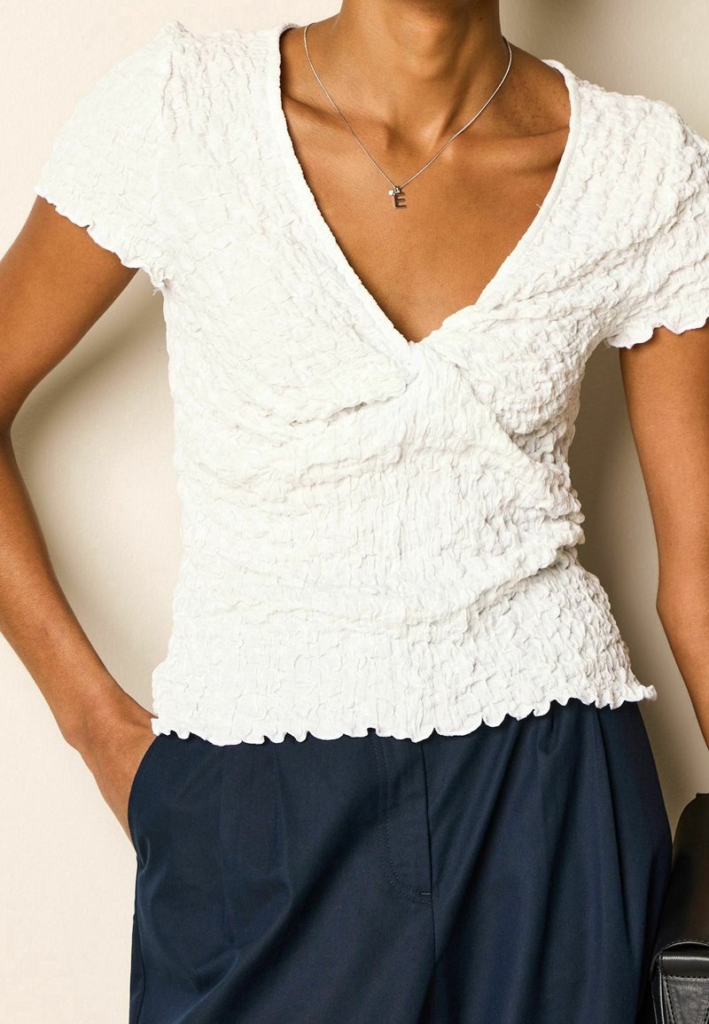 Personne portant un haut blanc texturé à manches courtes avec un col en V et un pantalon bleu marine, avec une main dans la poche et un collier en argent avec un pendentif en "E".