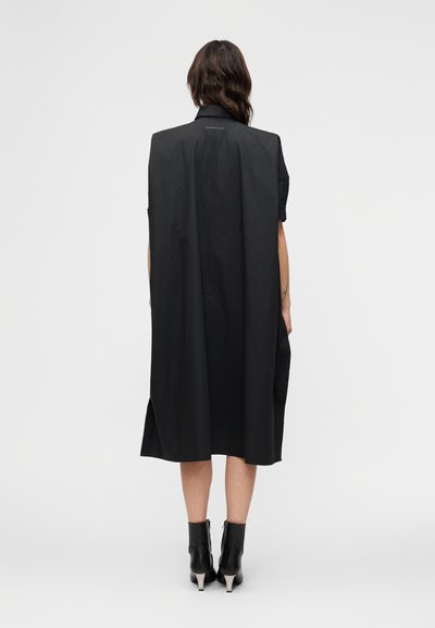 Γυναίκα με μακριά καστανά μαλλιά φορεί χαλαρό μαύρο παλτό μέχρι τα γόνατα με κοντά μανίκια και μαύρα μποτάκια με τακούνι, στέκεται σε λευκό φόντο.