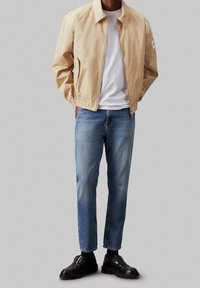 Ljusbeige bomberjacka med ståkrage, elastisk midja, fickor med dragkedja, bärs över en vit t-shirt och blå jeans, samt svarta skor.