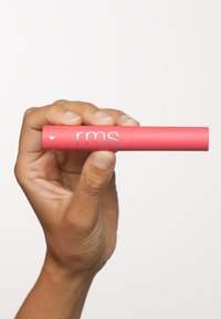 Rosa cylindrisk läppprodukt hålls i en hand, med silverlogotypen "rms" och uppåtpekande pilar på en slät mattyta.