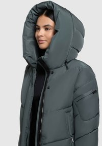 Vložna sive zimska jakna z velikim kapuco, zadrgo na sprednji strani, sprednjimi žepi in dekorativnim stranskim žepom; gladka tekstura.