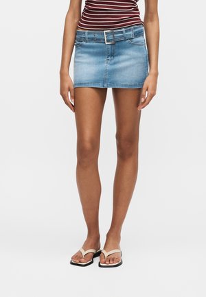 Vrouw draagt een lichtblauwe denim minirok met brede riem, gestreepte wijnrode top en beige sandalen met vierkante neus, staand tegen een witte achtergrond.