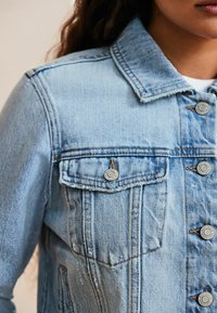 Chaqueta de mezclilla azul clara con cuello clásico, dos bolsillos frontales, botones plateados y detalles de costura visibles.