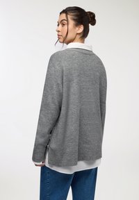 Maglione grigio a maglia con una vestibilità comoda, colletto rotondo e spacchi laterali, indossato sopra una camicia bianca, abbinato a jeans blu.