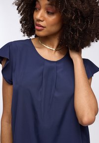 Top blu navy con maniche corte a volant, scollatura rotonda e dettagli a pieghe sottili. Tessuto liscio con una consistenza morbida. Collana choker dorata semplice.
