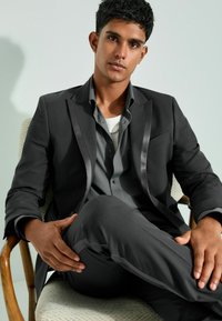 Traje negro con acabados de satén, camisa de botones gris, pantalones ajustados y una superficie texturizada, usados de manera casual mientras está sentado en una silla de madera.