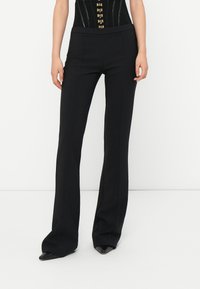 Woman wearing black high-waist, flared pants paired with pointed black shoes, standing against a plain white background.