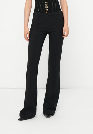 Woman wearing black high-waist, flared pants paired with pointed black shoes, standing against a plain white background.
