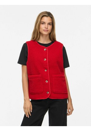 Jeune femme portant une chemise et un pantalon noirs, associés à un gilet rouge vif boutonné avec deux poches avant, debout devant un fond blanc.