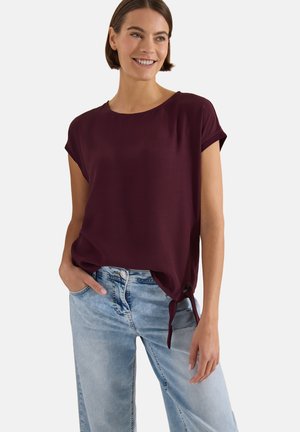 Kvinde smiler iført en kortærmet bourgognebluse med snøre detalje i siden og lyseblå slidte jeans, stående med den ene hånd i lommen.