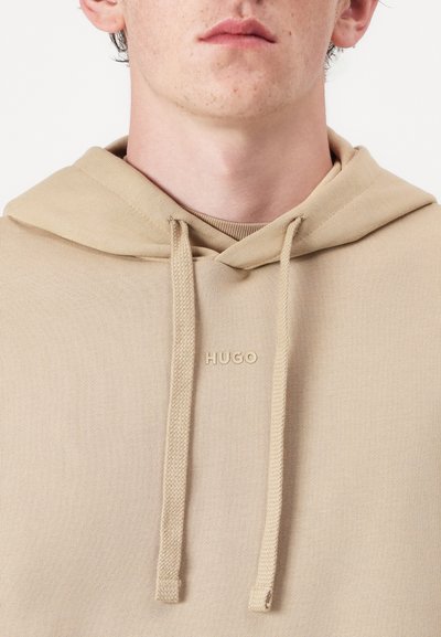 Hoodie beige avec capuche à cordon, tissu texturé et un petit logo en relief dans une teinte plus claire sur la poitrine.