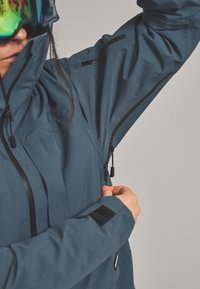 Blue waterproof jacket with a high collar, zippered vents, and velcro accents. Features a smooth texture and secure fastenings for functionality.