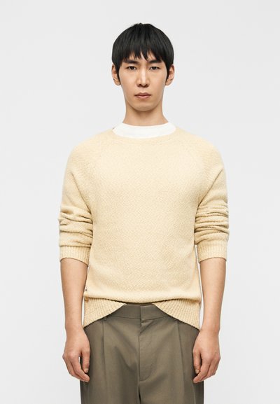 Maglione lavorato a maglia color crema con motivo testurizzato, polsini a coste e maniche raglan, abbinato a pantaloni chiari e larghi.