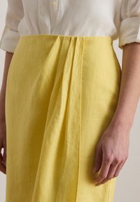 Hvit langermet skjorte stukket inn i høytlivs gul skjørt med plisserte detaljer, vist fra midjen til midt på låret på en stående person.