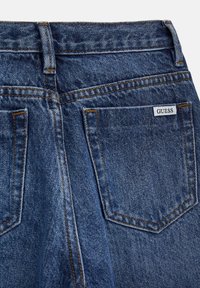 Jeans in denim di un blu medio con cuciture a vista e due tasche posteriori. Presenta un piccolo etichetta 'GUESS' vicino alla tasca destra.