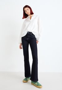 Vaqueros acampanados de mezclilla oscura con cintura alta, combinados con una camisa blanca de botones y zapatillas verdes con detalles amarillos.