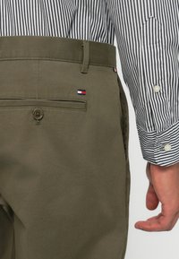 Olive green trousers with a smooth finish, featuring a button detail and a small red, white, and blue logo near the back pocket. Striped shirt visible.