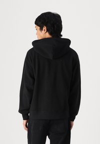 Hanorac din polar negru, cu un exterior texturat, buzunar tip kangaroo, glugă cu șnur și manșete și tiv din bumbac ribbit. Modelul este întors cu spatele.