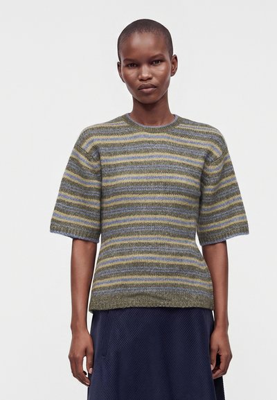 Personne aux cheveux courts portant un pull en tricot à manches courtes rayé olive, bleu et jaune, et une jupe bleu foncé.