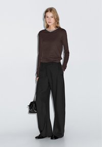 Bruine lange mouw shirt gelaagd over een grijze top, gecombineerd met zwarte wijde broek en een kleine zwarte handtas. Gladde, zachte texturen.