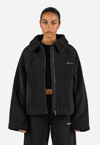 Giacca in pile nera con un taglio ampio, colletto largo, chiusura con zip e dettaglio del logo ricamato sul petto. Tessuto morbido e maniche oversize.