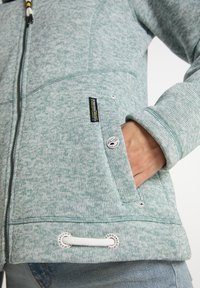 Ljusgrön texturerad fleecejacka med dragkedja framtill och fickor. Har tryckknappar, dragsko och logotyper på sidan.