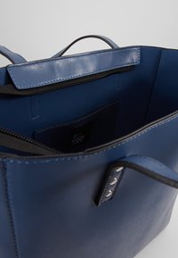 Wnętrze niebieskiej torebki wykonanej z syntetycznej skóry, z kieszenią na zamek, gładką teksturą oraz kontrastującymi czarnymi wykończeniami i metalowymi akcentami.