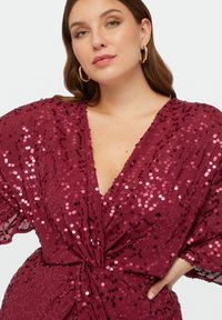 Robe en sequins bordeaux avec un décolleté en V profond, des manches chauve-souris courtes et un détail de taille noué, présentant une surface brillante et texturée.