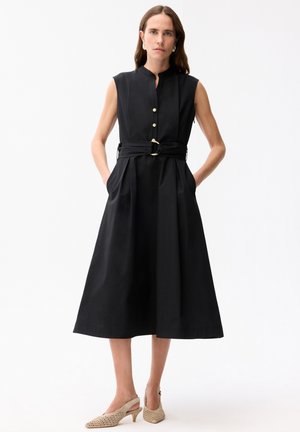 Vrouw staat met handen in zakken, draagt een mouwloze zwarte midi-jurk met gouden knopen en riem, gecombineerd met beige geperforeerde hakken.