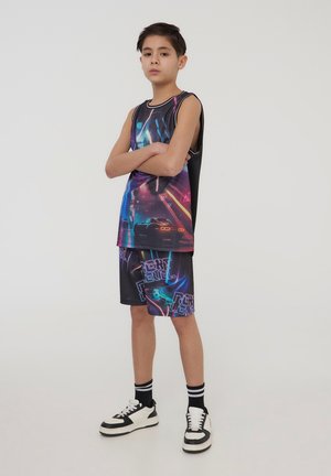 Canotta grafica nera con design di auto neon e accenti colorati, abbinata a pantaloni corti coordinati con loghi. Sneakers bianche e nere.