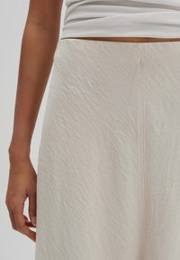 Jupe beige clair en tissu texturé avec un motif froissé. Présente un détail de couture, portée avec un haut blanc ajusté.