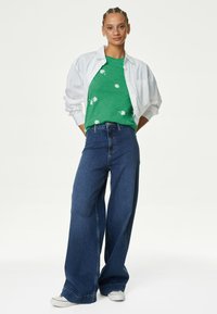 Camiseta verde con bordado floral, camisa blanca de gran tamaño y pantalones vaqueros de mezclilla oscuros de pierna ancha. La modelo lleva zapatillas plateadas.