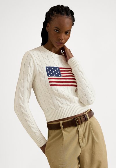 Bela pulover iz pletenega bombaža s cable knit vzorcem z vezeno rdečo in modro ameriško zastavo na sprednji strani. Model ga nosi s kaki hlačami in rjavim pasom.