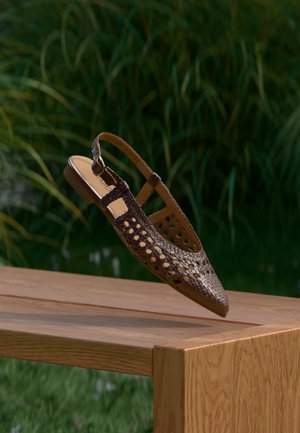 Bruine geweven slingback schoen met een puntige neus, voorzien van uitsnijdingen en een platte zool, geplaatst op een houten oppervlak.