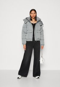 Grå pufferjakke med høj krave og aftagelig hætte, præget af vandret syning og to forlommer. Båret over en sort top og sat sammen med bredbenede jeans.