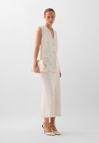 Roomkleurige tweed jurk met een mouwloze, met knopen sluitende top en bijpassende rok. Heeft een gestructureerd patroon en franjes aan de randen. Houdt een beige clutch vast.