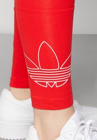 Rode leggings met een strakke pasvorm, voorzien van een wit Adidas-logo ontwerp op de kuit, gecombineerd met witte sportschoenen.